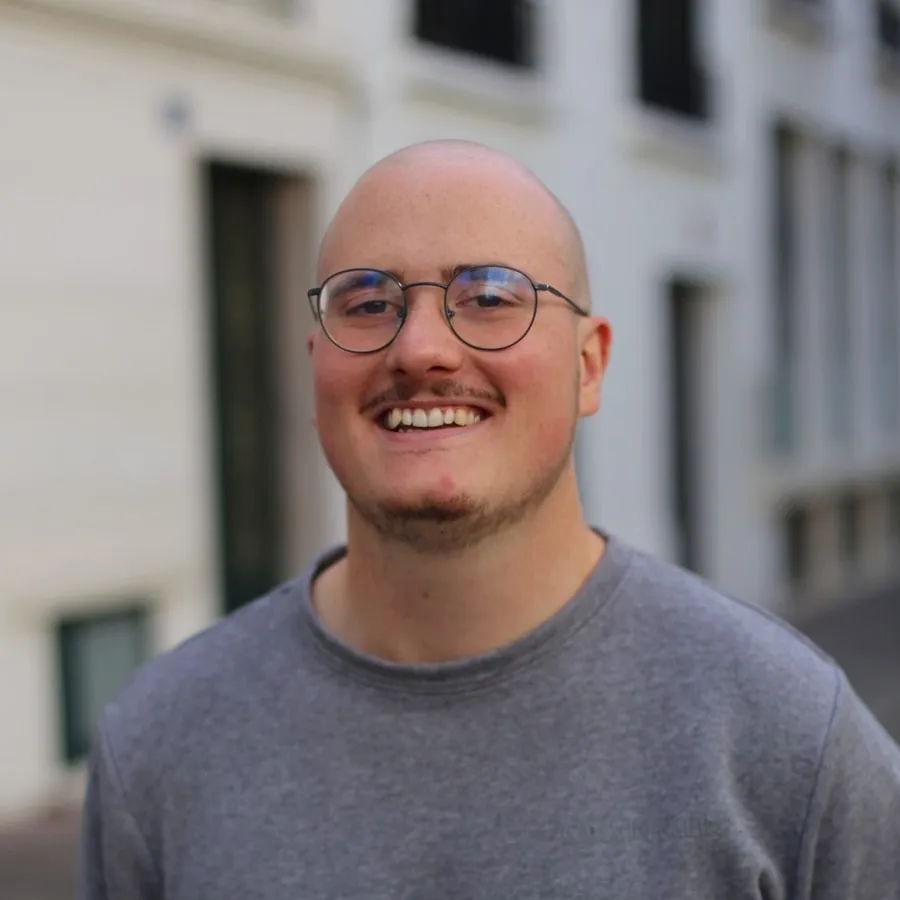 Portrait de Dylan Tracz, souriant, avec des lunettes rondes et un pull gris, dans une rue.